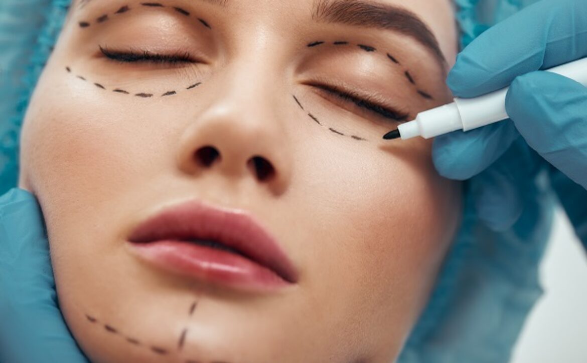 Becoming perfect. Close up photo of young woman with glowing skin in blue medical hat waiting for facial surgery while plastic surgeon in blue gloves drawing dashed line under eyes